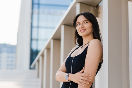 A young woman in black sportswear poses near the wall of a modern building. She is wearing earbuds and looks confident.の写真素材