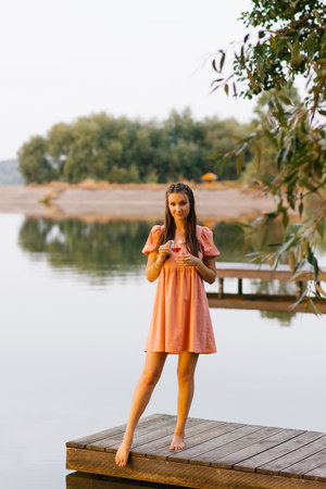A woman in a peach colored dress stands barefoot on a wooden pier, with a serene lake view in the background.の写真素材