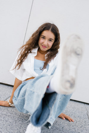 A playful girl from Generation Alpha sitting on the ground, extending her leg forward to highlight her sneakers, smiling confidently.の写真素材