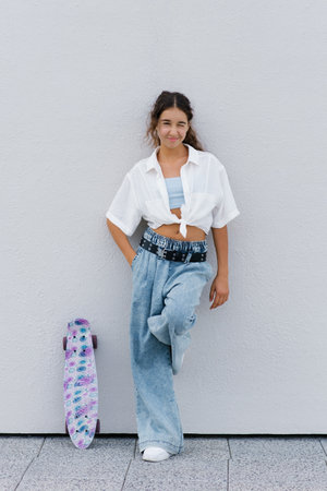 A Gen Alpha girl posing against a white wall with her colorful skateboard, expressing playful and relaxed emotions.の写真素材