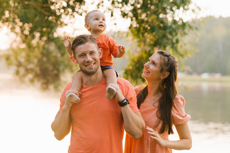 Happy family enjoying a sunset outdoors near a lake. The father carries the child on his shoulders, while the mother smiles warmly. The golden hour lighting creates a soft, peaceful atmosphere.の写真素材
