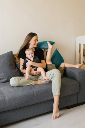 A mother sits on a gray sofa holding a baby, while her older child playfully jumps with a pillow in a cozy, bright room.の写真素材