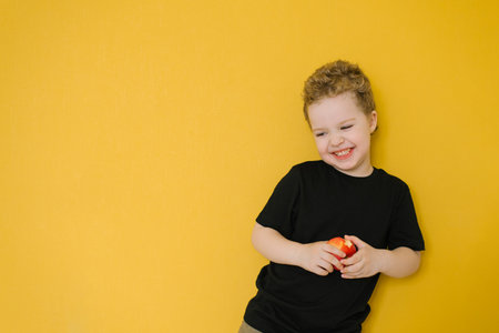 A young boy in a black t shirt holding a red apple, standing against a bright yellow wall, smiling...