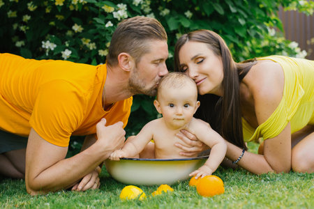 A family enjoying a loving moment outdoors, with the parents kissing their baby who is sitting in a small tub surrounded by oranges. The scene is filled with warmth, joy, and affection.の写真素材