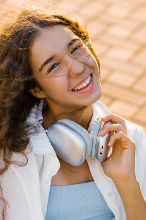 A joyful girl with a radiant smile and curly hair, holding silver headphones under golden sunlight....