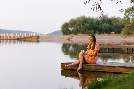 A woman in a peach dress sits gracefully on a wooden pier with a tea cup in her hand, reflecting on the serene water around her.の写真素材