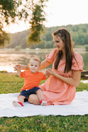 A mother and her toddler sitting together on a blanket near a lake during sunset. The golden light...