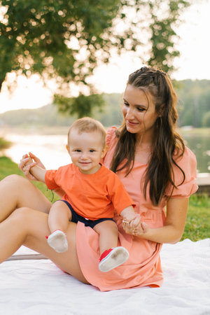 A baby sitting on his mother lap and smiling, surrounded by a serene picnic setup by the lake, with...