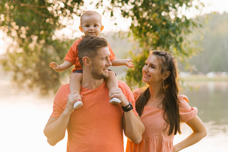 Happy family enjoying a sunset outdoors near a lake. The father carries the child on his shoulders, while the mother smiles warmly. The golden hour lighting creates a soft, peaceful atmosphere.の写真素材