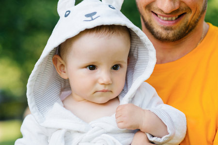 Close up of a baby wearing a white hooded robe, looking curious and calm while enjoying a green park environment.の写真素材