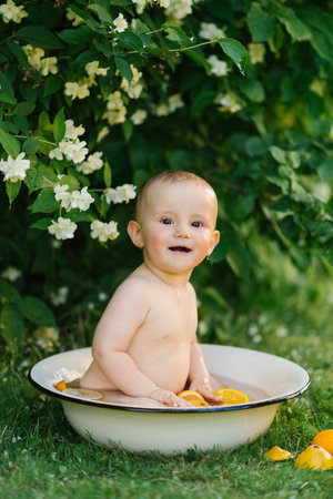 A happy baby sitting in a small tub outdoors, surrounded by water and oranges, with a big smile on...
