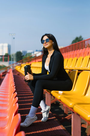 Woman in black sportswear sitting on yellow stadium seats, holding a black shaker bottle and enjoying a sunny day.の写真素材