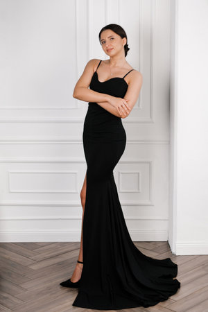 A young woman in a sleek black evening gown poses gracefully against a white paneled wall, showcasing elegance and timeless beauty in a minimalistic interior setting.の写真素材