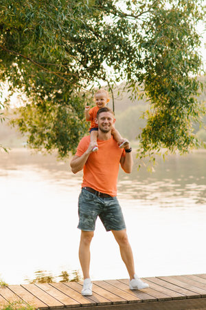A father holds his toddler son on his shoulders, standing near a serene lake at sunset. Both are smiling, enjoying the peaceful outdoor environment together.の写真素材