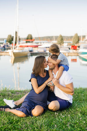 A pregnant mother sits with her family near a calm marina, enjoying a tranquil summer day with boats and greenery in the background.の写真素材