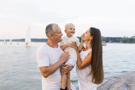 Happy family of three, man and woman and baby, are standing on pier by the water