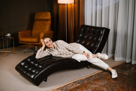 A woman sitting comfortably in a black leather chair, smiling in a warm and stylish psychotherapist's office with brown tones.の写真素材