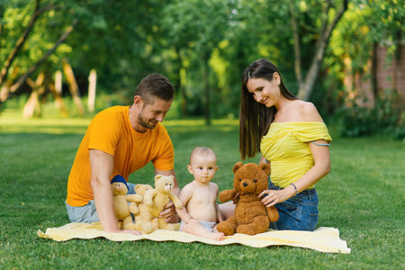 A family sitting together on a yellow blanket outdoors, playing with teddy bears, with the baby...