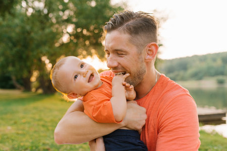 A father lovingly holds his baby boy while sitting on a white blanket in a park during sunset, creating a warm and joyful family moment.の写真素材
