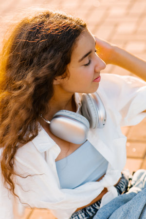 A teenage girl with curly hair sitting outdoors in warm sunlight, holding silver headphones, with a thoughtful and calm expression.の写真素材