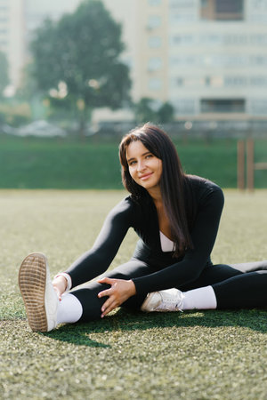 A young woman in black sportswear stretching her legs on a grass field under the bright sun,...