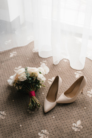 A romantic wedding bouquet and white shoes placed on a patterned carpet with soft light coming through the curtains.の写真素材