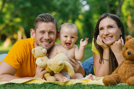 A happy family spending time outdoors with teddy bears, a joyful baby playing and laughing between the parents.の写真素材