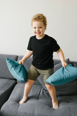 Young boy holding two blue cushions and standing on a gray sofa, smiling joyfully.の写真素材