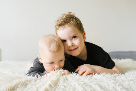 Older sibling gently leaning on a baby brother, lying on a fluffy white rug, showing casing affection and warmth.の写真素材