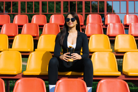 A woman in black sportswear sitting on bright yellow and red stadium seats, holding a shaker bottle...
