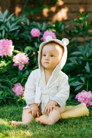 A baby wearing a white robe with bear ears, sitting in a garden surrounded by blooming pink flowers on a sunny day.の写真素材