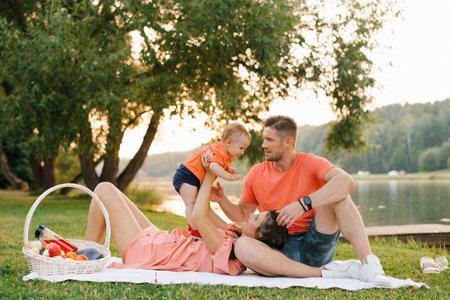A happy family enjoying quality time together by a lake during a summer picnic. Parents and toddler dressed in orange outfits, surrounded by nature beauty.の写真素材