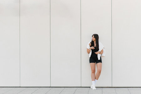 Full body portrait of a young woman in a black sports outfit with a white sweatshirt, smiling confidently against a white background outdoors.の写真素材