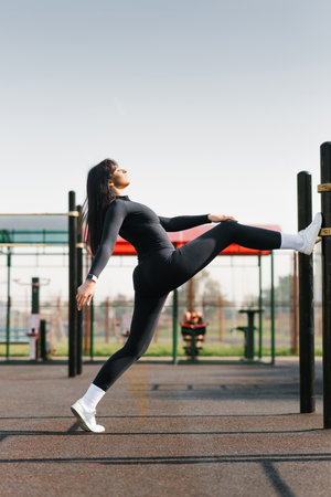 A young woman in black sportswear performing a stretching exercise on outdoor fitness equipment under a clear blue sky.の写真素材