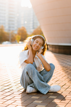 A teenage girl sitting on a brick-paved surface in casual denim and a white shirt, wearing large headphones and smiling in soft evening sunlight.の写真素材