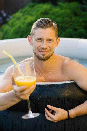 A man sitting in an outdoor pool, holding a glass of orange juice with a straw, surrounded by lush greenery in the background.の写真素材