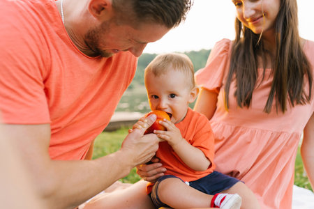 A father helping his toddler eat a fresh peach while sitting on a blanket near the lake. The family enjoys a relaxing picnic surrounded by nature.の写真素材