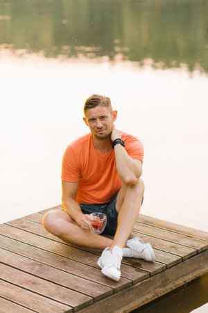 A cheerful man in an orange shirt sits on a wooden pier, enjoying tea and the scenic view of a lake during a summer evening.の写真素材