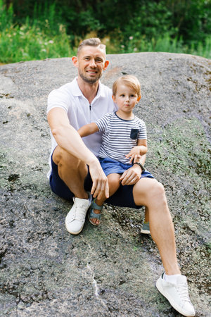 A smiling father sitting with his son on a large rock outdoors, enjoying a peaceful and joyful moment in nature.の写真素材