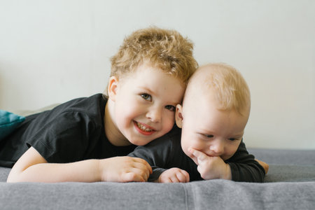 Two brothers, a baby and a toddler, lying on a gray sofa, smiling warmly and enjoying each other's...