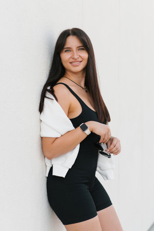 A portrait of a Caucasian young woman in her twenties outdoors, dressed in a white sweatshirt over her shoulders and a black outfit, smiling against a white wall.の写真素材