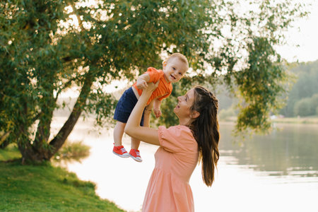 A loving mother holding her baby near a serene lake with vibrant green surroundings and the warm...
