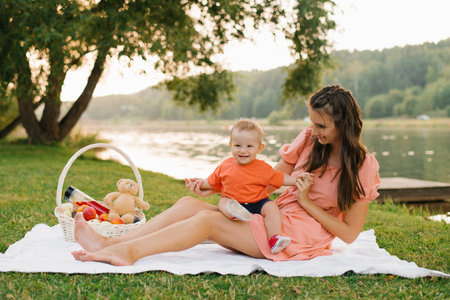 A joyful baby sitting on his mother lap during a picnic by the lake, with a basket of fruits and...