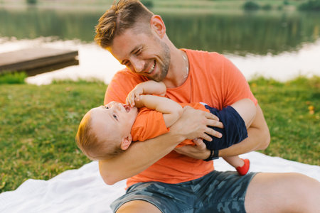 A father enjoys playful moments with his baby boy while sitting on a picnic blanket in a scenic park...