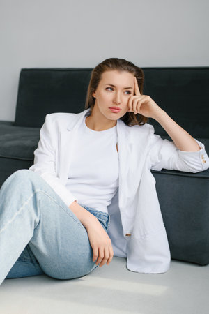 Thoughtful young woman in a white blazer and jeans sitting on the floor by a dark sofa in a bright minimalist interior, ideal for lifestyle, fashion and business content.の写真素材