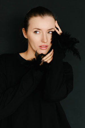 Studio portrait of a young woman in a black sweater with feather details, posing against a dark background, ideal for fashion, beauty, and editorial concepts.の写真素材