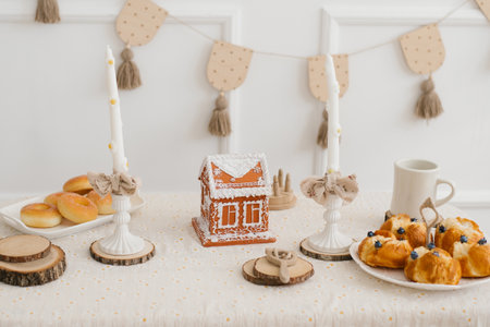 Cozy festive table setting with a gingerbread house, fresh pastries, and elegant candles on a light...
