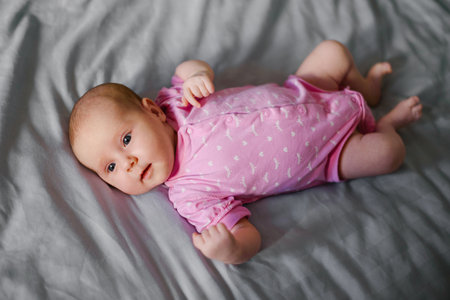 Newborn baby in pink onesie lying on gray bedding, gazing to the side with a calm and curious...