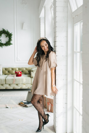 Stylish young woman in beige dress standing near a bright window and smiling, with festive home decor in the background. Perfect for lifestyle, holiday, and fashion related visuals.の写真素材