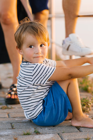 Barefoot boy in striped shirt and blue shorts sitting on a wooden pier by the lake at sunset, looking to the side. Ideal for summer lifestyle, childhood, and outdoor leisure themes.の写真素材